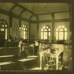 2009 Archives Week Postcard, an image of the Guilford College Library c. 1909. Two people sitting at a table in a large room with other tables and charis and a desk with someone sitting at it. 4 tall windows surrounding the room with a high ceiling.