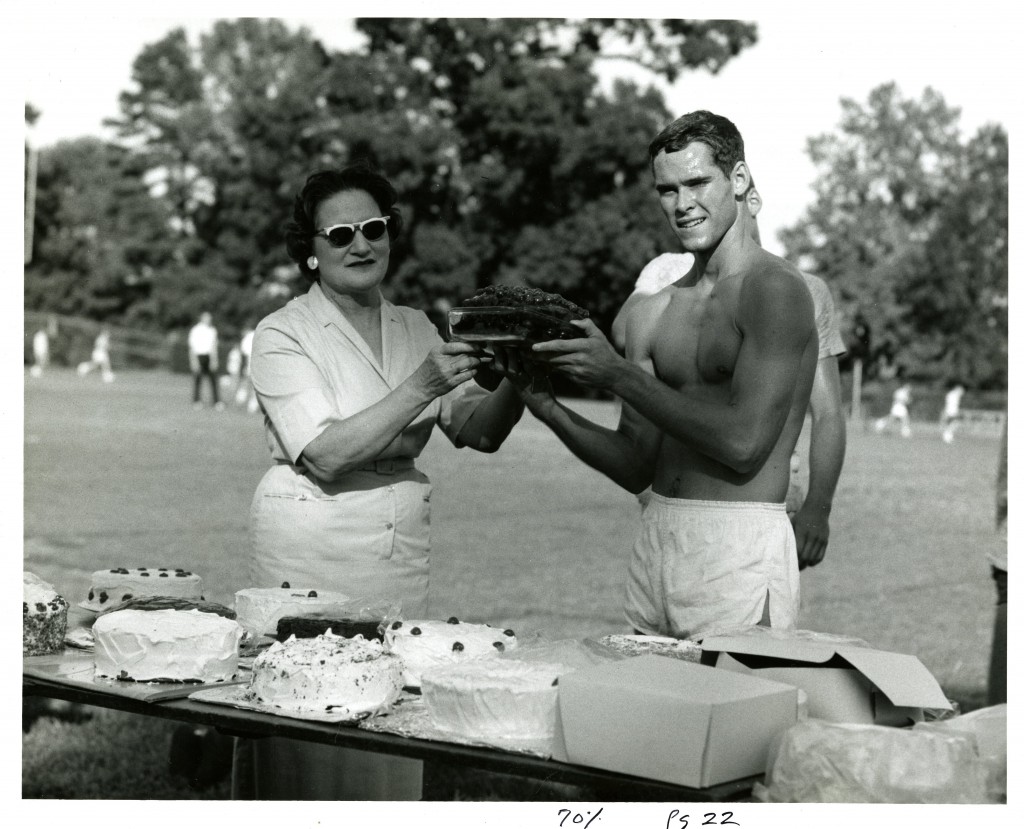 Daisy Whittle presents a cake to a winning racer(who isn't wearing a shirts) in 1963, while there is a table full of cakes right next to them and people running the the blurred background