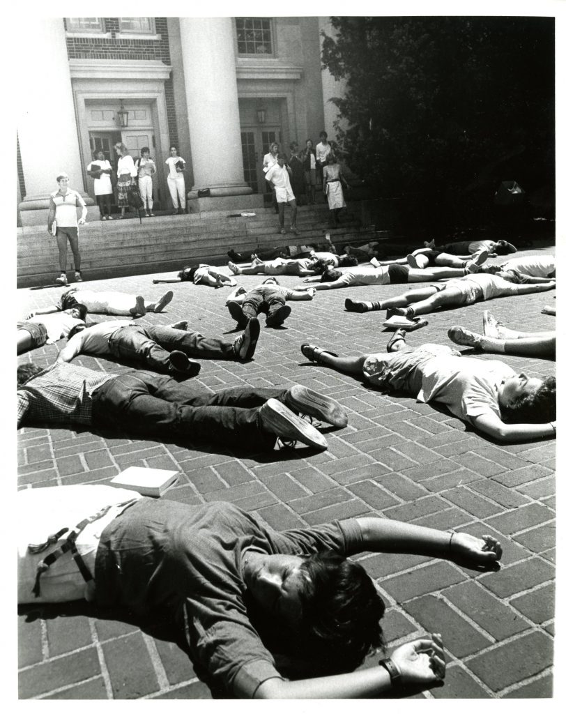 A photo capturing students participating in the die-in inf front of Chambers Building. Students lying on the ground appearing to be dead outside of chambers while other students are walking out of chambers