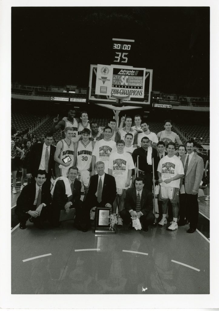 Team picture of the 1998 men's basketball team after winning the Southern Conference Tournament on the court with the hoop right behind them and a sign saying, "1998 Champions"
