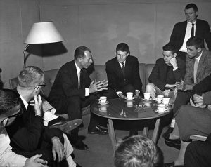 Dean Burts engaging with students, everyone is wearing a suit and drinking out of teacups all around a small round table sitting on a couch