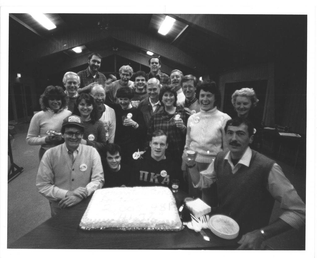 Davidson employees gather around a cake with icing spelling out "Congratulations Davidson, 2,007,481, 41.7%" at a Development retreat in 1986. Bill is seated far right, next to the cake.