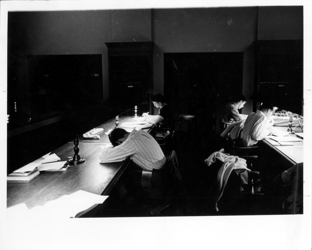 A student sleeping in the Grey Library reading room while two others study, circa 1960.