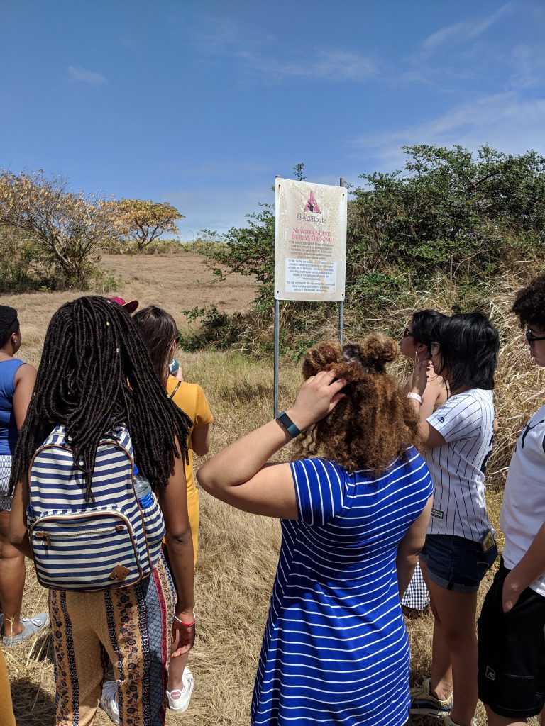 Several students gather around a plaque in a large field marking the cemetery of enslaved persons in Barbados.